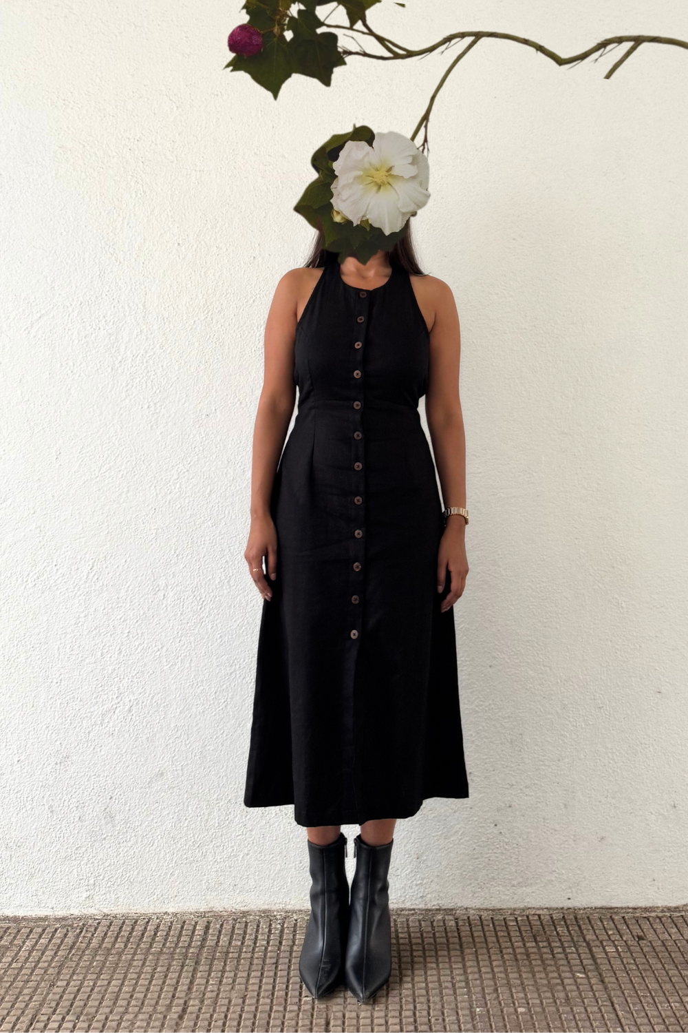 A person wearing a sleeveless black midi dress with wooden buttons, paired with black boots, standing against a white background with a large flower partially covering the face.