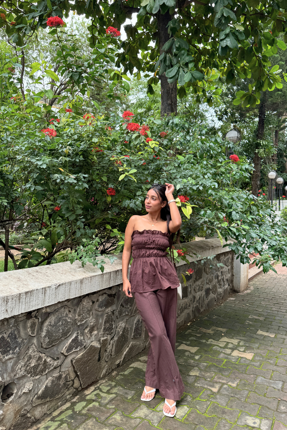 Woman in a BROWN outfit standing in a garden with greenery and flowers.